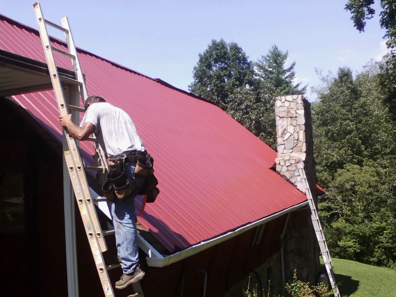 Roofer on a ladder beside a red metal roof and stone chimney — Soffit & Fascia Repair by Roofing Brothers in Cottontown
